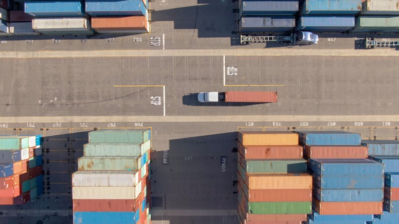 White truck transports a red freight container across the busy port of Los Angeles. Flying above a freight truck hauling a heavy shipment past large stacks of other containers. 